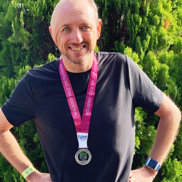 Participant Paul Bovington happily smiling after achieving his running goal – he is wearing a silver medallion with a pink satin ribbon around his neck proudly.