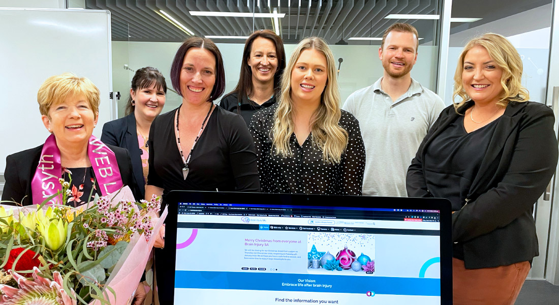 Website Team happliy standing in a group together with the new website in the foreground displayed on imac computer